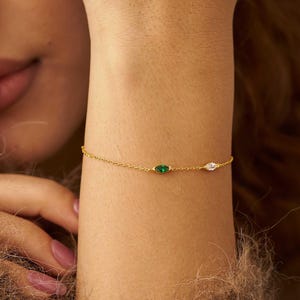 May include: A delicate gold bracelet featuring two oval-shaped gemstones: a green emerald and a clear, sparkling stone. The bracelet is worn on a wrist, showcasing its fine chain and elegant design. The jewelry is a subtle yet eye-catching accessory.