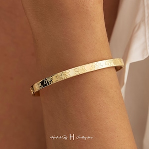 May include: A gold-toned metal bangle bracelet featuring a repeating engraved floral pattern along its outer surface. The bracelet has a smooth, polished finish and a slim, uniform band design that encircles the wrist with a simple, clean aesthetic.