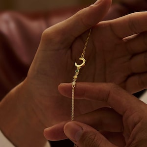 May include: A gold-toned chain bracelet featuring a small crescent moon charm followed by three tiny circular gemstones in green, yellow, and clear white. The delicate metal chain includes an adjustable extender section at the end of the bracelet.