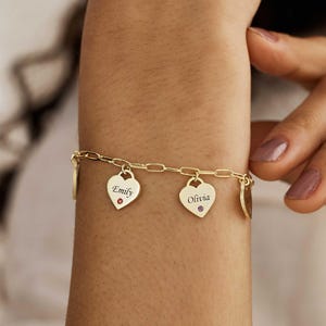 May include: A gold-toned chain link bracelet featuring multiple heart-shaped charms. Two of the charms are engraved with the names Emily and Olivia, each accented with a small, singular coloured gemstone at the base of the heart engraving.
