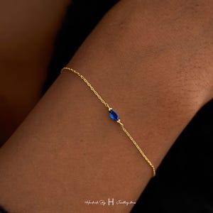 May include: A delicate gold chain bracelet featuring a single marquise-cut blue gemstone centered on the thin metal link strand. The jewelry piece is designed with a minimalist aesthetic, showcasing a vibrant sapphire-colored stone set in gold.