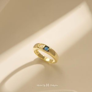 Könnte beinhalten: Ein goldener Bandring mit zwei kleinen eingesetzten Steinen: einem klaren, dreieckigen Stein und einem quadratisch geschliffenen blauen Stein. Der Ring hat eine glatte, polierte Oberfläche und ein schlichtes, abgerundetes Design. Der Text "Handmade By H Jewellery store" ist sichtbar.