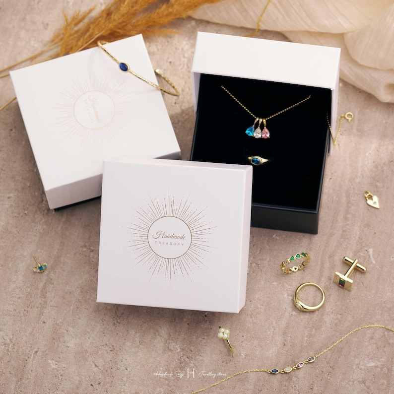 May include: A collection of gold jewelry including a delicate bracelet, a necklace with three pear-shaped pendants, several rings, a pair of cufflinks, and a heart-shaped charm, displayed alongside two white gift boxes labeled Handmade Treasury.