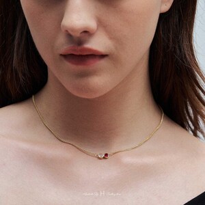 May include: A delicate gold chain necklace featuring two small, circular bezel-set stones. One stone is a clear, translucent white, and the other is a vibrant, deep red. The necklace rests against the neck, showcasing a simple, refined design.