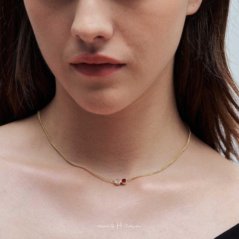 May include: A delicate gold chain necklace featuring two small, circular bezel-set stones. One stone is a clear, translucent white, and the other is a vibrant, deep red. The necklace rests against the neck, showcasing a simple, refined design.