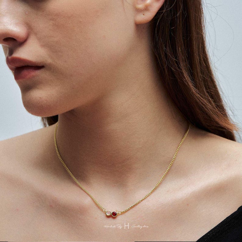 May include: A gold-toned chain necklace featuring two small, circular charms positioned off-center. One charm is a clear, translucent stone, and the other is a deep red, circular stone. The necklace rests against the neck with a delicate link design.