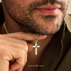 May include: A gold cross pendant necklace featuring a diagonal row of small blue stones across the center and a small bail set with tiny clear stones, suspended from a thin gold chain. The text Handmade By H Jewellery stones is visible.