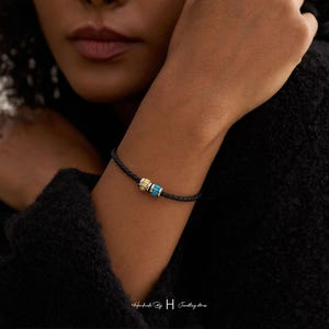 May include: A black braided cord bracelet featuring two small cylindrical beads. One bead is gold-toned with a textured surface, and the other bead is bright blue with a metallic finish. The bracelet is worn around the wrist.