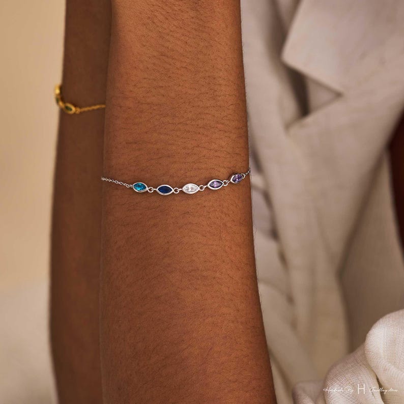 May include: A delicate silver chain bracelet featuring five marquise-cut gemstones in a row. The stones display a gradient of colors including light blue, dark blue, clear white, light purple, and a deeper purple, all set in polished silver metal links.