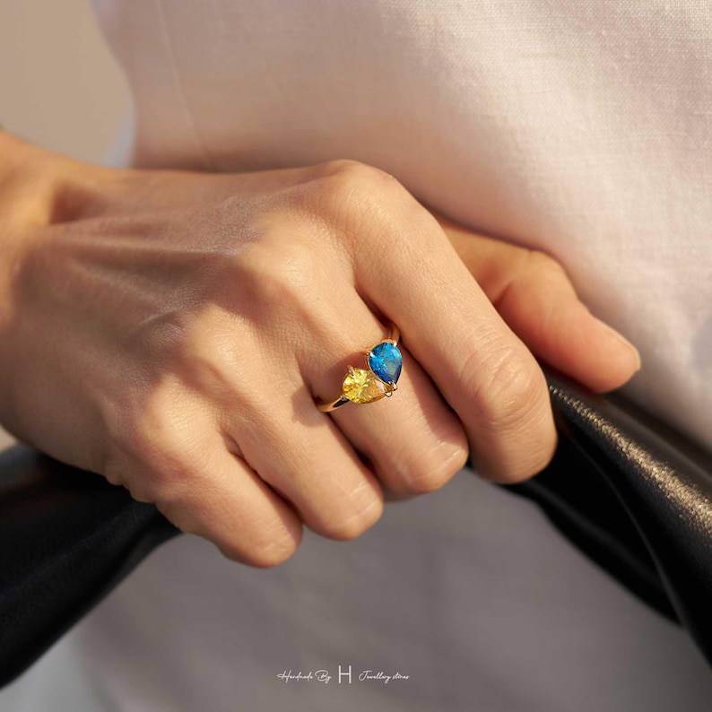 May include: A gold ring featuring two pear-shaped gemstones, one in a vibrant blue and the other in a bright yellow, set side-by-side on a thin gold band. The design highlights the contrasting colors of the teardrop-shaped stones in a simple setting.