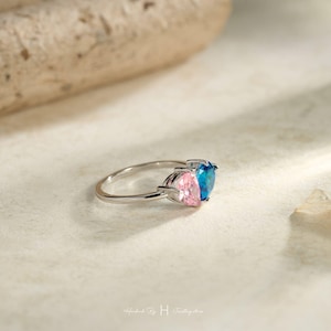May include: A silver-toned ring featuring two heart-shaped gemstones set side-by-side. One gemstone is a vibrant blue, and the other is a soft pink. The band is thin and metallic, showcasing a polished finish that reflects light across its surface.