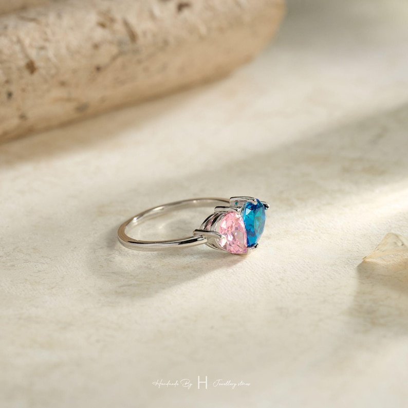 May include: A silver-toned ring featuring two heart-shaped gemstones set side-by-side. One gemstone is a vibrant blue, and the other is a soft pink. The band is thin and metallic, showcasing a polished finish that reflects light across its surface.