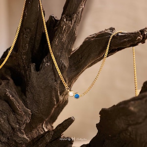 May include: A gold-toned chain necklace featuring two small, circular bezel-set stones, one clear and one deep blue, suspended from the center. The delicate metal chain links are uniform in size and color, creating a simple, refined jewelry piece.