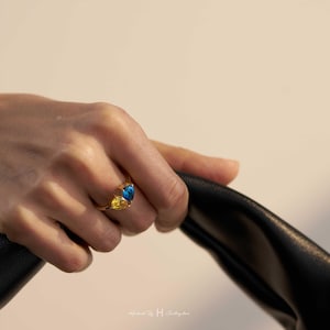 May include: A gold ring featuring two pear-cut gemstones, one in a vibrant blue and the other in a bright yellow, set side-by-side on a thin metallic band. The design highlights the contrasting colors of the stones in a simple, elegant arrangement.