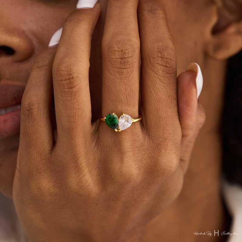 May include: A gold ring featuring two pear-shaped stones set side-by-side. One stone is a deep green color, and the other is a clear, colorless stone. The stones are held in place by gold prongs on a thin, polished gold band.