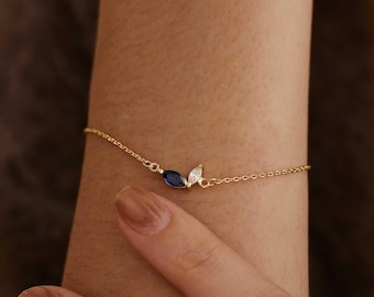 May include: A delicate gold chain bracelet featuring two marquise-cut stones, one deep blue and one clear white, arranged in a leaf-like pattern. The bracelet is secured with a thin, polished gold link chain that wraps around the wrist.