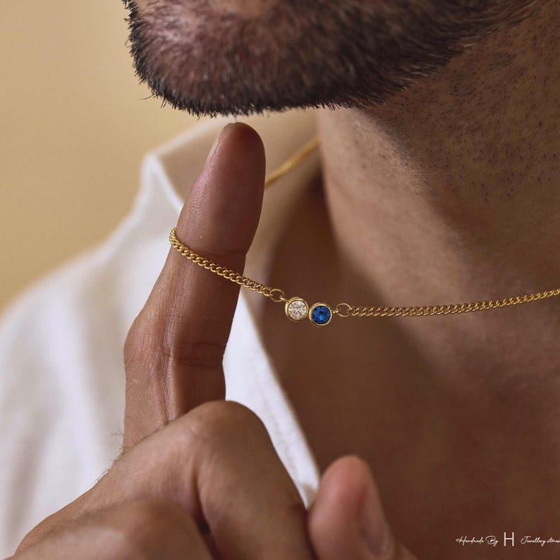 May include: A gold chain necklace featuring two small, circular bezel-set stones: one clear white gemstone and one vibrant blue gemstone. The delicate metal chain connects to each stone setting, creating a simple and refined jewelry piece.