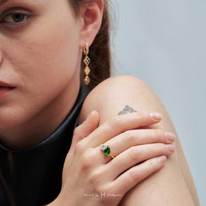 May include: A gold ring featuring a pear-cut clear stone and a pear-cut green stone set side-by-side on a thin band, paired with a gold drop earring consisting of three connected diamond-shaped links with black and clear stone accents.