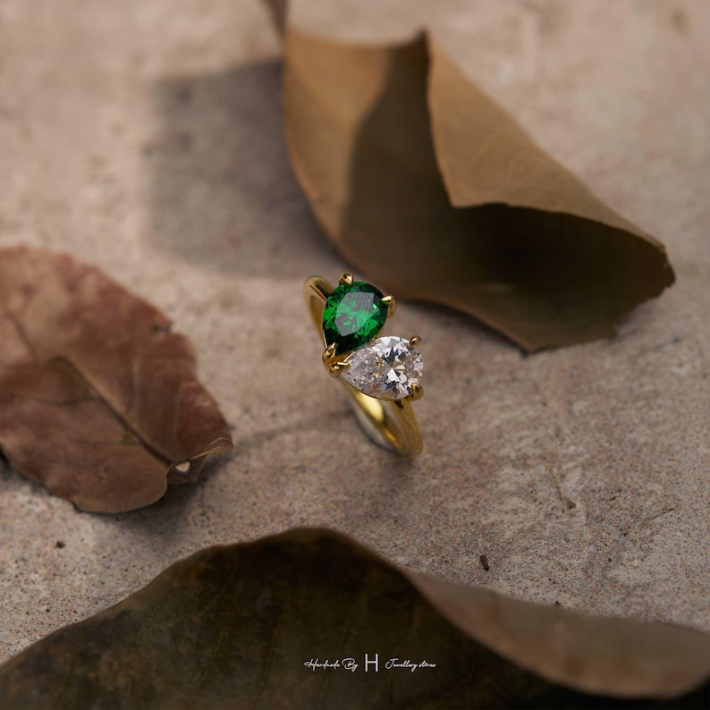 May include: A gold ring featuring two pear-cut stones, one vibrant green and one clear white, set in a bypass style. The band is polished gold metal. The text "Handmade By H Jewellery store" is printed in small white letters at the bottom.