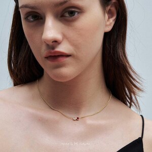 May include: A delicate gold chain necklace featuring two small, circular charms. One charm is a clear, colorless stone, and the other is a vibrant red stone. The necklace rests against the neck, showcasing a simple, refined design with a thin metal link chain.