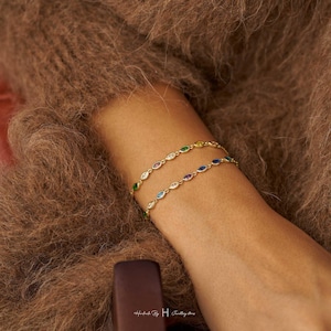 May include: Two gold chain bracelets featuring marquise-cut gemstones in various colors including green, blue, yellow, and purple. The bracelets are delicate and link together with small metal loops. The text Handmade by H Jewellery stores is visible.