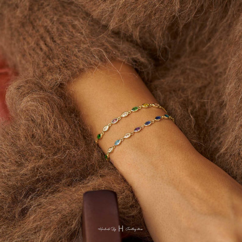 May include: Two gold chain bracelets featuring marquise-cut gemstones in various colors including green, blue, yellow, and purple. The bracelets are delicate and link together with small metal loops. The text Handmade by H Jewellery stores is visible.