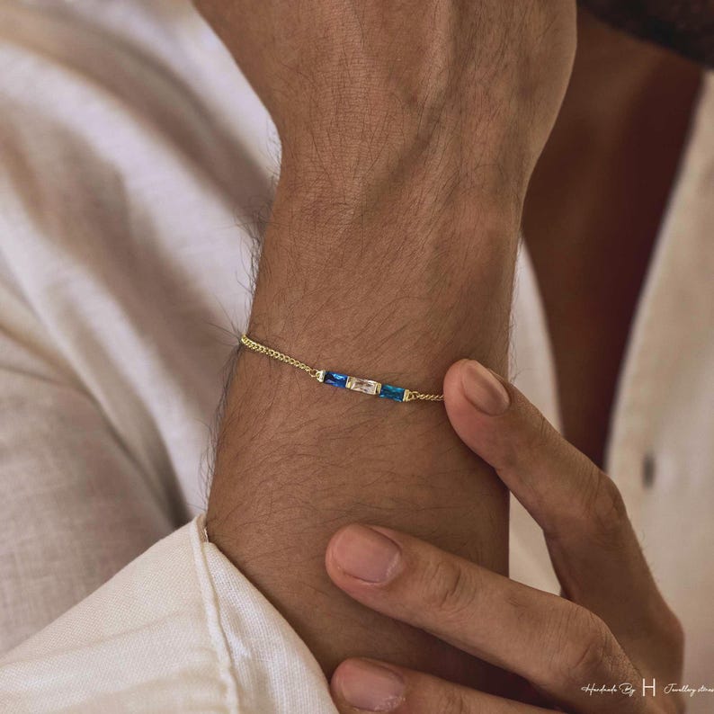 May include: A delicate gold chain bracelet featuring three rectangular baguette-cut stones arranged horizontally in the center. The two outer stones are deep blue, while the central stone is clear and colorless, set in a thin gold metal frame.