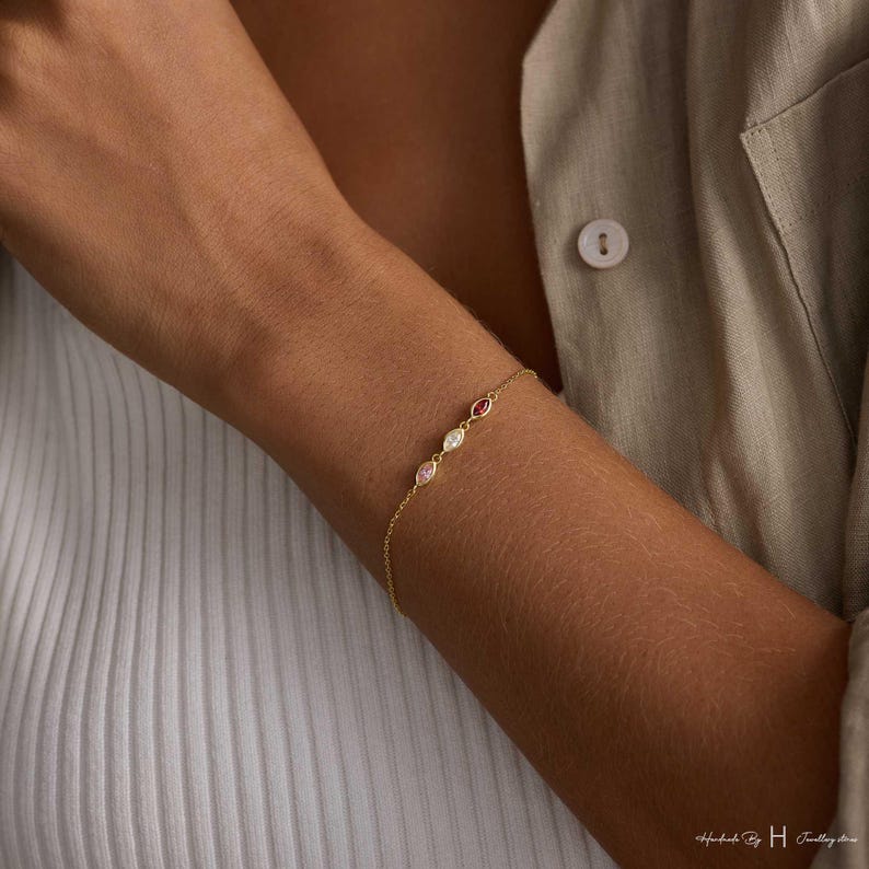 May include: A delicate gold chain bracelet featuring three marquise-cut gemstones in a row. The stones are set in gold and include a pink, a clear, and a red gemstone. The bracelet is secured with a thin gold chain that wraps around the wrist.