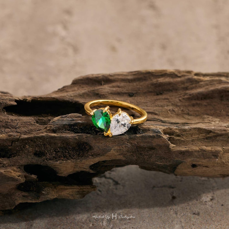 May include: A gold ring featuring two pear-cut stones set side-by-side, one in a vibrant emerald green and the other in a clear white, creating a dual-stone design on a polished gold band that rests on a piece of textured dark brown wood.