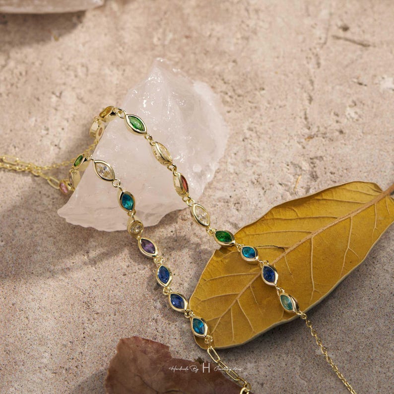 May include: A gold-toned chain bracelet featuring a series of marquise-cut, multi-colored gemstones in shades of green, blue, purple, and clear white. The delicate metal links connect each stone, creating a continuous, slender, and elegant jewelry piece.