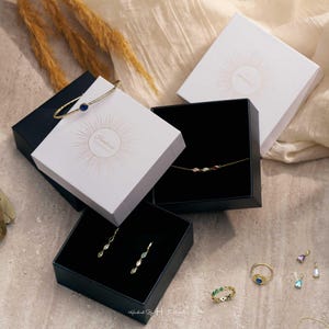 May include: A collection of gold-toned jewelry including a thin bangle with a blue stone, a delicate chain necklace with multi-colored stones, drop earrings, and several rings with colorful accents, all presented in white and black gift boxes.