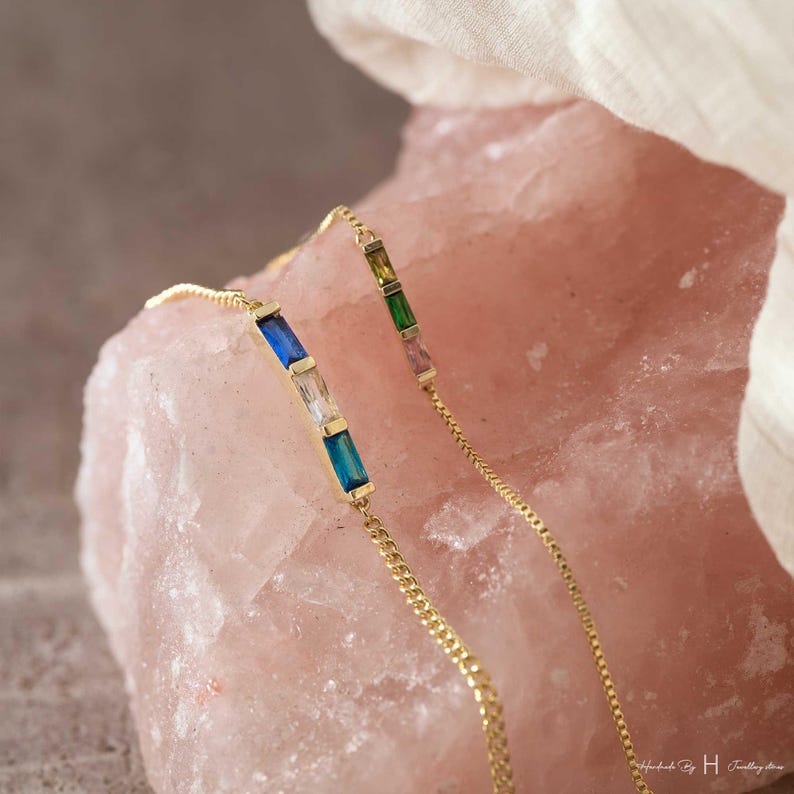 May include: Two delicate gold chain bracelets featuring baguette-cut gemstones. One bracelet displays a blue, clear, and blue stone sequence, while the other shows a green, yellow, and pink stone arrangement. Both chains are thin and metallic.