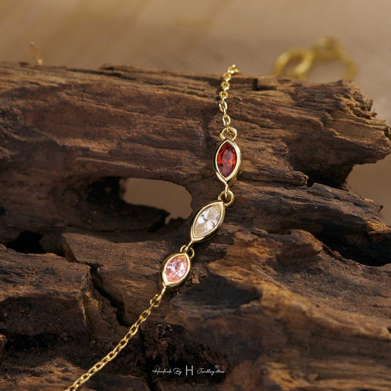 May include: A delicate gold chain bracelet featuring three marquise-cut gemstones in red, clear, and light pink. The bracelet is composed of thin gold links connecting each stone setting. The text "Handmade By H Jewellery Store" is visible.