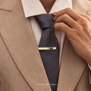 May include: A gold-toned tie bar featuring three small square-cut gemstones in clear, red, and green, secured across a dark grey diagonal-striped necktie worn with a white collared shirt and a tan blazer. The text Handmade By H Jewellery stones is visible.