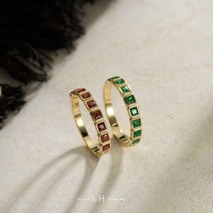 May include: Two gold rings featuring a row of square-cut gemstones. One ring is set with deep red stones, while the other ring is set with vibrant green stones. Both bands have a polished gold finish and a channel setting for the square stones.