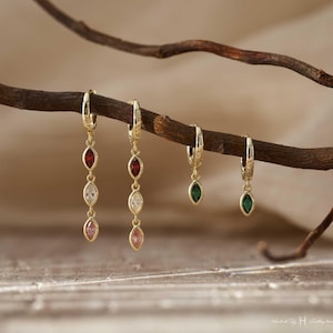 Op de afbeelding: Gouden oorringen met kleurrijke marquise-geslepen edelstenen. Twee paar oorbellen hebben bungelende edelstenen in rood, wit en roze. De andere twee paar hebben groene edelstenen. De oorbellen worden op een bruine tak getoond.