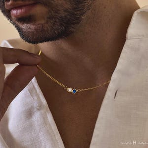 May include: A gold chain necklace featuring two small circular bezel-set stones, one clear white and one deep blue, positioned side-by-side. The delicate metal chain connects to each side of the stone settings, creating a simple and refined jewelry piece.