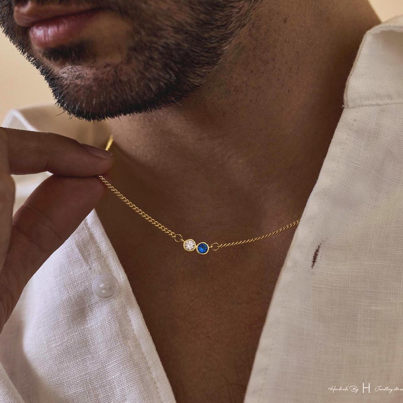 May include: A gold chain necklace featuring two small circular bezel-set stones, one clear white and one deep blue, positioned side-by-side. The delicate metal chain connects to each side of the stone settings, creating a simple and refined jewelry piece.