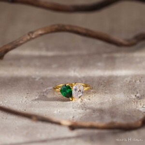 May include: A gold ring featuring two pear-cut stones set side-by-side. One stone is a deep emerald green, and the other is a clear, colorless stone. The band is thin and metallic gold, creating a dual-stone design that sits prominently on the surface.
