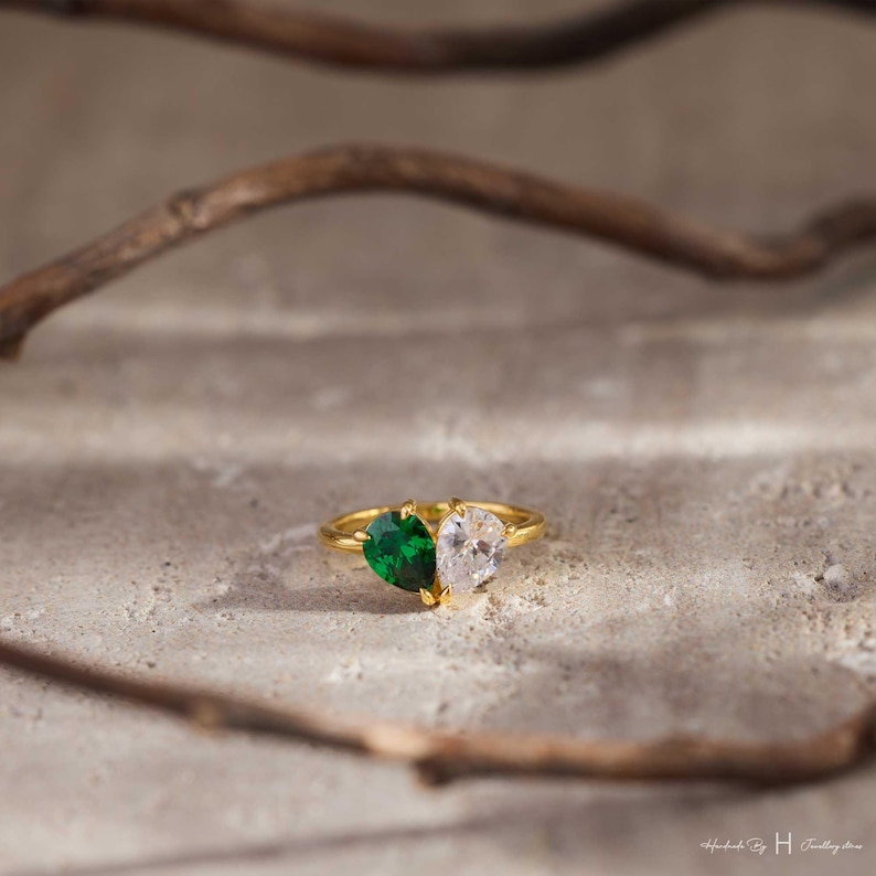 May include: A gold ring featuring two pear-cut stones set side-by-side. One stone is a deep emerald green, and the other is a clear, colorless stone. The band is thin and metallic gold, creating a dual-stone design that sits prominently on the surface.