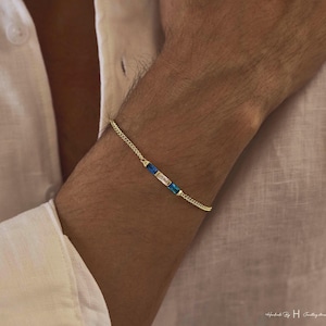 May include: A delicate gold chain bracelet featuring three rectangular baguette-cut stones arranged in a row. The center stone is clear, flanked by two vibrant blue stones on either side, all set within a thin, polished gold-tone metal link chain.