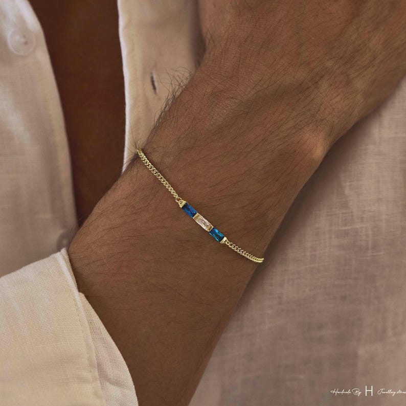 May include: A delicate gold chain bracelet featuring three rectangular baguette-cut stones arranged in a row. The center stone is clear, flanked by two vibrant blue stones on either side, all set within a thin, polished gold-tone metal link chain.