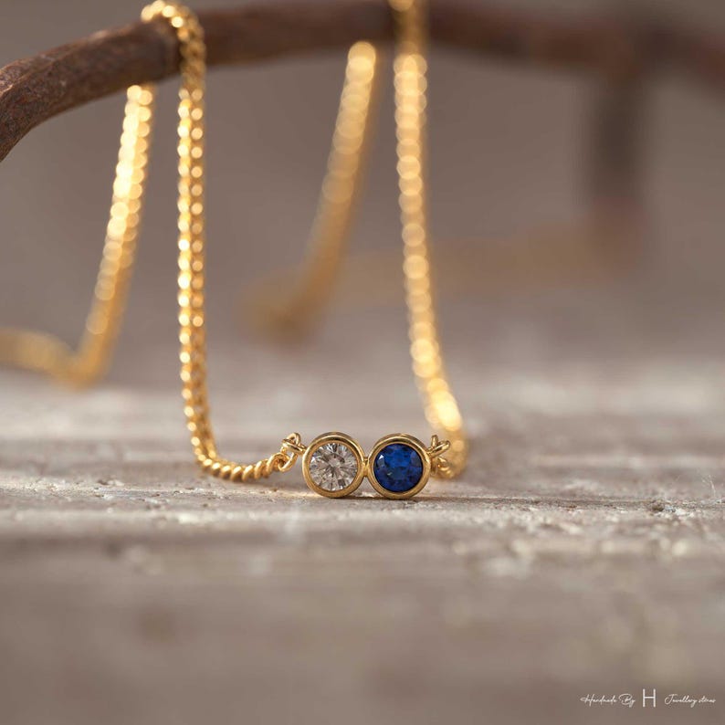May include: A gold chain necklace featuring two circular bezel-set stones side-by-side. One stone is a clear, faceted white gemstone, and the other is a deep blue, smooth-surfaced gemstone. The necklace is draped over a curved brown branch.