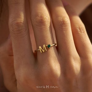 May include: A gold-toned ring featuring a capital letter M and a small, dark green heart-shaped gemstone. The band is thin and polished, with the letter and stone positioned side-by-side on the top of the ring, creating a delicate, open design.