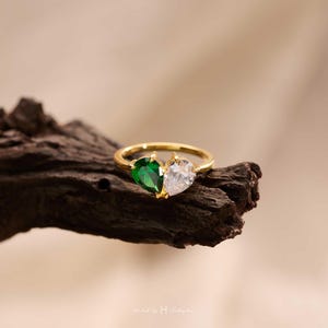 May include: A gold ring featuring two pear-cut stones set side-by-side to form a heart shape. One stone is a deep emerald green, and the other is a clear, colorless stone. The band is a thin, polished gold metal that encircles the finger.