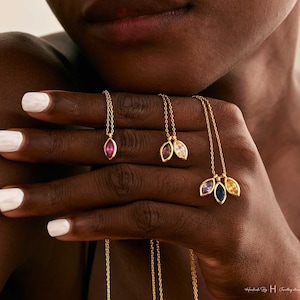 May include: Three gold necklaces featuring marquise-cut gemstones. One necklace has a single pink stone, one has two stones in red and clear, and one has three stones in purple, blue, and yellow. The chains are thin gold links.