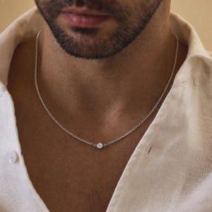 May include: A silver-toned chain necklace featuring a single, small, round, clear stone set in a bezel setting at the center. The necklace rests against the neck, showcasing a delicate link design that connects to the central stone setting.