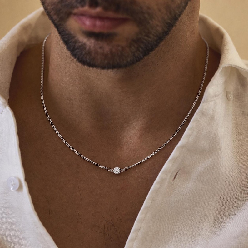 May include: A silver-toned chain necklace featuring a single, small, round, clear stone set in a bezel setting at the center. The necklace rests against the neck, showcasing a delicate link design that connects to the central stone setting.