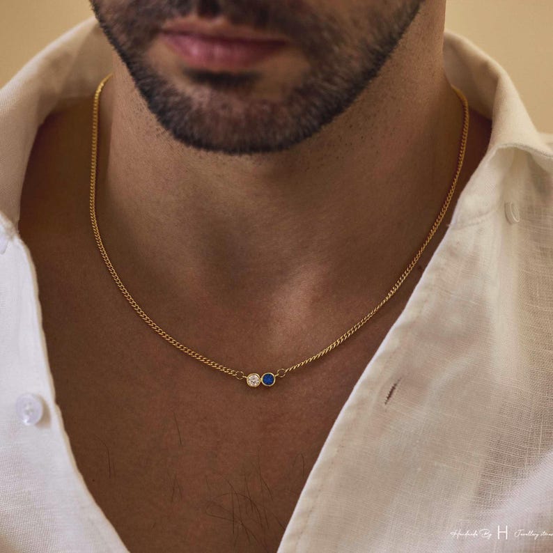 May include: A gold chain necklace featuring two small circular pendants positioned at the center. One pendant contains a clear white gemstone, while the adjacent pendant holds a deep blue gemstone. The necklace is worn against a white collared shirt.