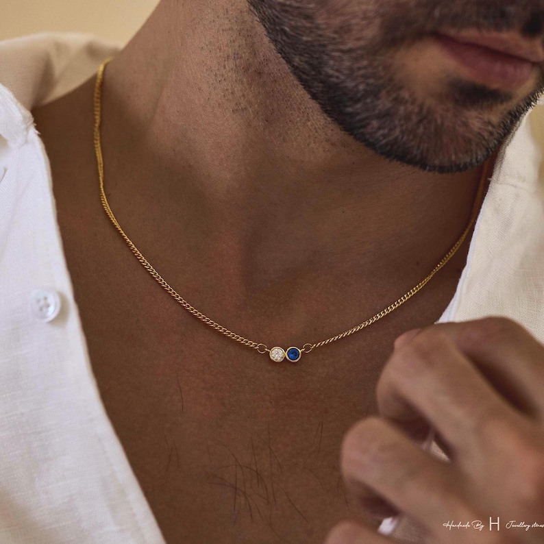 May include: A gold chain necklace featuring two small circular pendants, one with a clear white stone and the other with a deep blue stone, resting against the neck. The necklace is worn with a white collared shirt.